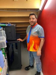 Austin, a smiling, dark haired young man stands with his hand on a large computer network server which he has helped install.